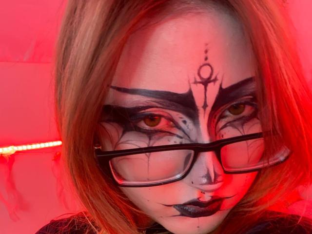ShainaBallensky with red makeup, black eyeliner, black lipstick, nose ring, and glasses, has a black face tattoo above her eyes, under red lighting, with blonde hair.
