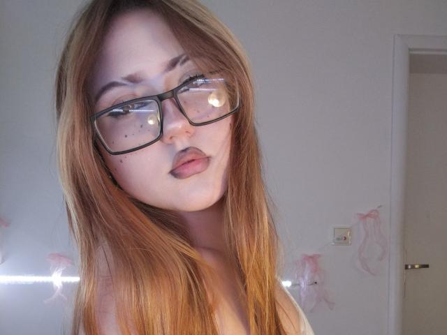 ShainaBallensky with long, straight, auburn hair, wearing black-framed glasses, has freckles on her nose, and is looking at the camera with a slight pout, showing her bare shoulders.
