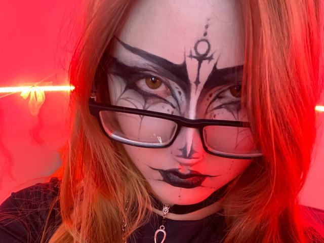 ShainaBallensky with goth makeup, black lipstick, and heavy black eyeshadow, wearing glasses and a black choker, has red hair and a nose ring, against a red-lit background.