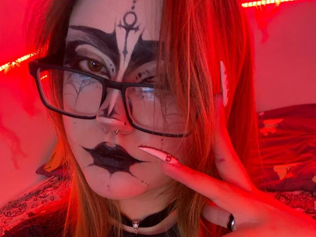 ShainaBallensky with red-tinted lighting, black makeup, glasses, nose ring, black lipstick, finger on lip, choker, and tattoos, has long orange hair and a pierced nose.