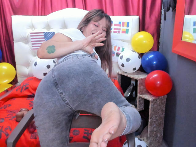 SofiaXDelRio deitada em uma cadeira com as pernas levantadas, vestindo calças jeans cinzas, mostrando uma tatuagem no braço superior, e com o pé levantado, cercada por balões coloridos..