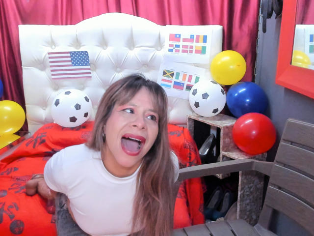 SofiaXDelRio, com longos cabelos castanhos, está ajoelhada em uma cama, com a boca aberta em uma expressão de surpresa, vestindo uma camisa branca, cercada por bolas de futebol e balões coloridos..
