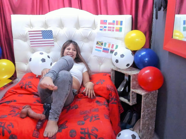 SofiaXDelRio, barefoot, lying on a red bed with soccer balls, flags, and balloons, wearing gray pants, white top, smiling, foot up, red curtain, wooden nightstand.
