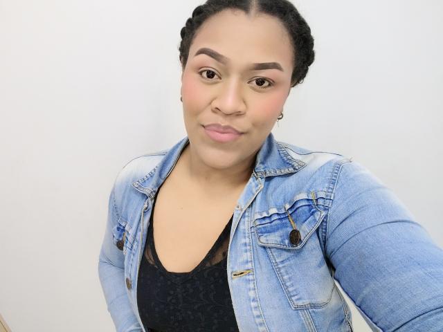 KloheEvans, com cabelo trançado, veste uma jaqueta jeans azul por cima de uma blusa preta de renda, tem um rosto fofo, levemente emburrado, e olhos castanhos pequenos e redondos. Seus seios mal são visíveis..