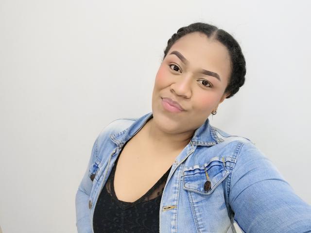 KloheEvans mit schwarzen Zöpfen, heller Hautfarbe, trägt eine Jeansjacke über einem schwarzen Spitzenoberteil, lächelt leicht, hat rosafarbene Lippen, braune Augen, kleine Ohrringe und ein dezentes Make-up..