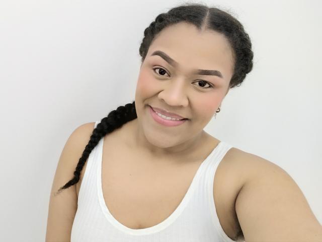 KloheEvans tem um sorriso adorável, pele marrom clara, cabelo preto trançado, uma camiseta branca e olhos castanhos. Ela está mostrando seus pequenos seios e lábios rosados..