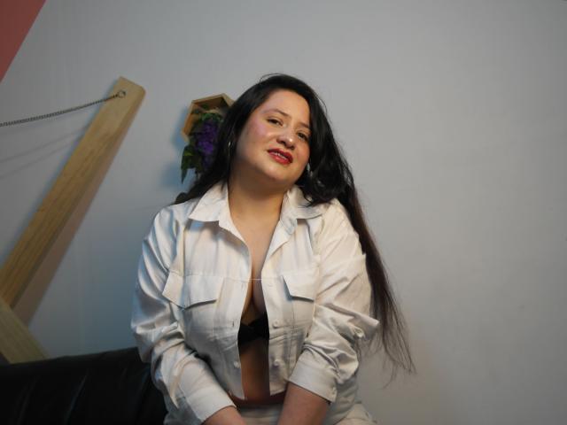 LauraCastilloECH tem cabelos pretos longos, batom vermelho e está sorrindo, vestindo uma camisa branca aberta que revela seu sutiã preto e seios pequenos. Ela está ligeiramente inclinada para frente..