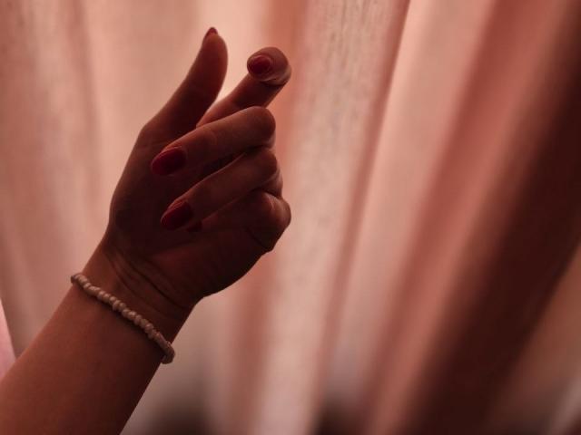 Rozalins Hand mit rot lackierten Nägeln und einem weißen Perlenarmband, leicht erhoben. Ein zarter, rosa Vorhang im Hintergrund, leicht unscharf. Ihre Finger sind anmutig gekrümmt..