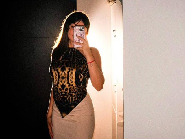 AshlyLee with long dark hair, wearing a leopard print halter top and tight beige skirt, standing in a mirror selfie, phone covering face, light behind her.