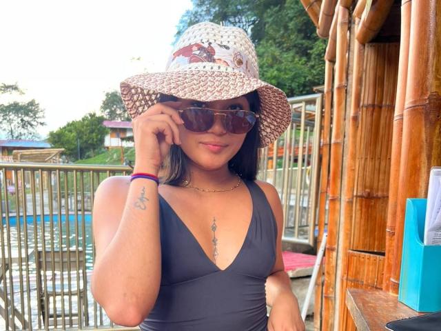 AshlyLee with tan skin, wearing a black one-piece, wide-brimmed straw hat, sunglasses, and a necklace, touching her hat, showing a tattoo below her boobs.