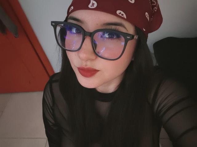 Susan Monroy, met zwarte bril, rode bandana en rode lippenstift, draagt een doorschijnend zwart topje, waardoor haar borsten lichtjes zichtbaar zijn door de stof. Ze zit met een zwarte tas achter zich..