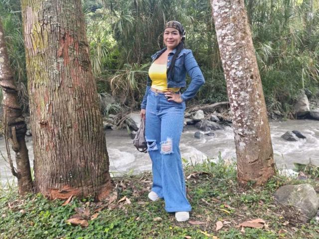 TamaraFoxy met een bruine huid, bruin haar, die een gele top, blauwe versleten jeans en een blauwe jas draagt, staat bij bomen, ziet er zelfverzekerd uit, met haar hand op haar heup, en er is een rivier op de achtergrond te zien..