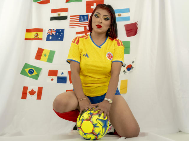 LizBonnet, in een hurkhouding, draagt een geel voetbalsok, blauwe korte broek, rode sokken, en houdt een geel en blauw voetbal vast. Op de achtergrond zijn kleurrijke vlaggen te zien. Ze heeft rode lippenstift op, een gemiddelde borstgrootte, bruine huid en zwart en rood haar..