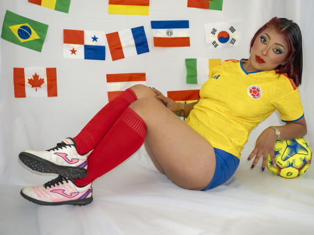 LizBonnet with yellow soccer shirt, red socks, blue shorts, and white soccer shoes, sitting with legs up, holding a soccer ball, wearing bold eye makeup and red lipstick.