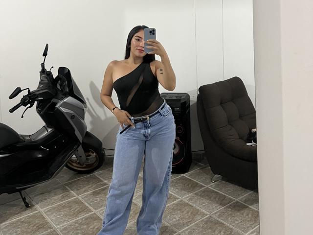 SharonKlart with one boob showing, wearing a black one-shoulder top, blue jeans, and a black phone, standing next to a scooter and a black chair.
