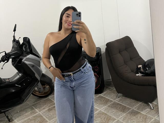SharonKlart with long black hair, wearing a black one-shoulder top, showing tits through sheer fabric, and blue jeans, holding a phone, standing in front of a scooter.