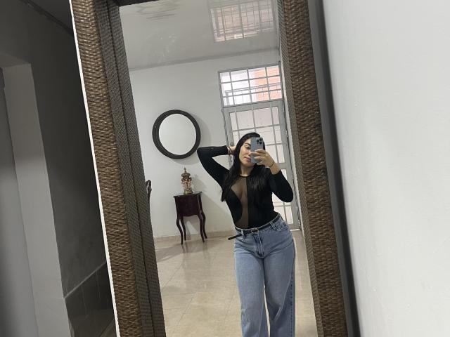 SharonKlart with long black hair, wearing a black sheer long-sleeve top and high-waisted blue jeans, taking a mirror selfie, showing her tits and ass.