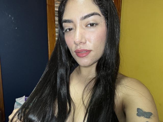 SharonKlart with long black hair, brown eyes, pink lips, nose piercing, and a butterfly tattoo on her right shoulder. She's wearing a sleeveless top, looking slightly annoyed.