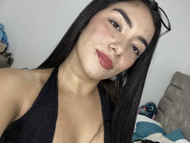 SharonKlart with long black hair, wearing a black halter top, glossy red lips, and eyeglasses on her head, smiling at the camera, showing her medium tits.