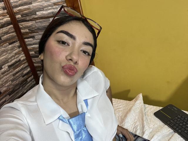 SharonKlart with puffy pink lips, wearing a white shirt and blue tie, has red-framed glasses on her head, sitting on a bed with a yellow wall behind her.