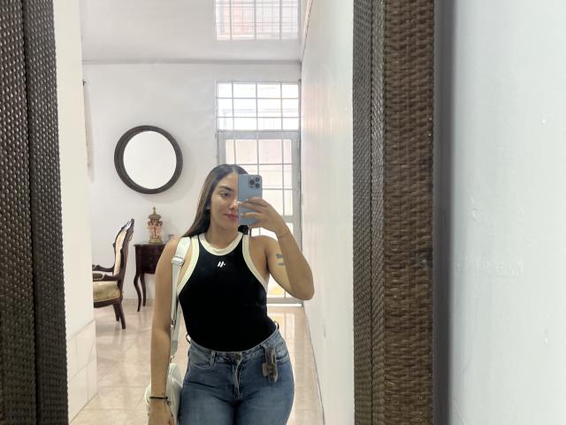 SharonKlart with long brown hair, black tank top, white straps, blue jeans, holding phone, mirror reflection, white walls, wicker frame, window, chair with yellow cushion, small tattoo.