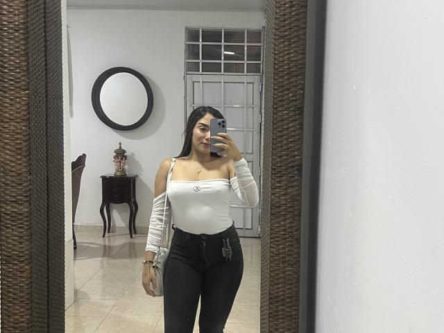 SharonKlart with long black hair, white off-shoulder top, black high-waisted pants, holding phone in mirror selfie, small tits, slim, standing in room with wicker mirror frame.