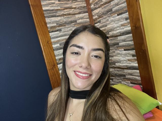 SharonKlart has long brown hair, a black choker, and is smiling with pink lips, showing off her shiny skin and small nose. Her tits are not visible.