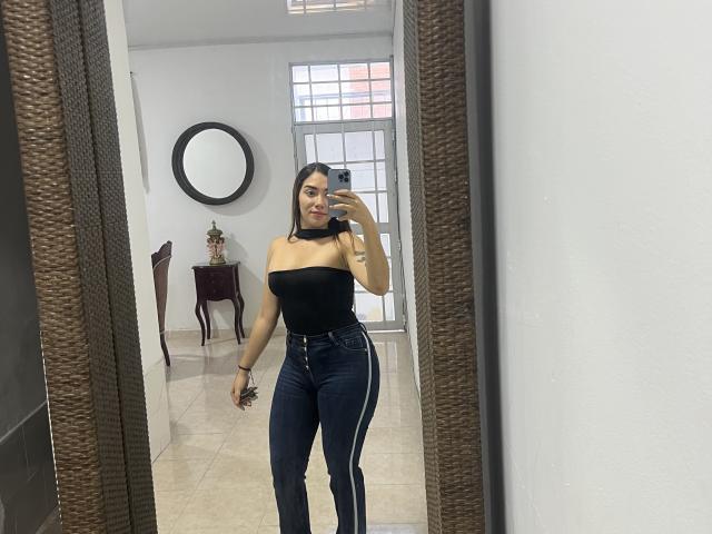 SharonKlart with black strapless top, showing her tits, and dark blue track pants with white lines, taking a mirror selfie, holding phone, in a room with white walls.
