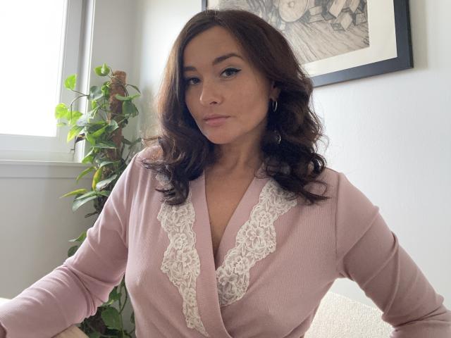 Dionisia with medium brown wavy hair, pink lace-trimmed top, showing her tits, sitting in a bright room, green plant in background, framed black-and-white artwork on white wall.