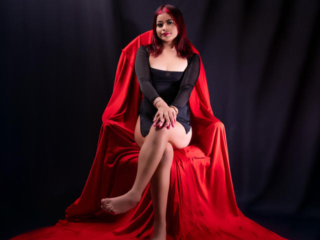 JennieRider with red hair, black dress, and red satin draped over throne, sitting with legs crossed, hands on knees, red nails, black background, looking at camera.