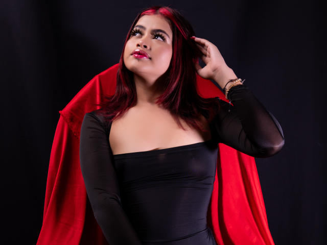 JennieRider with red hair, black dress, and red cape, hand in hair, looking up, pink lipstick, black background, wrist bracelet, medium tits, confident, sexy, stylish.