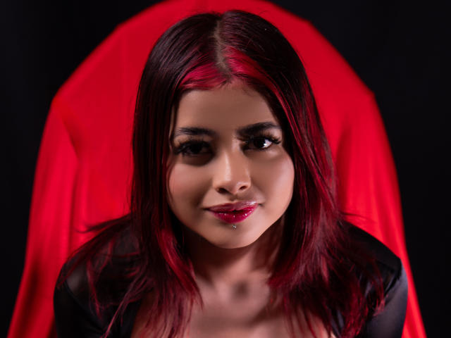 JennieRider has red and black hair, dark eyes, and glossy pink lips. She's wearing a black top and sitting against a red backdrop. Her small nose and pierced lip are visible.