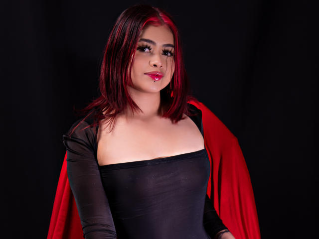 JennieRider with red hair, black sheer top, and red cape, showing her tits, pink lipstick, and a slight smile against a dark background.