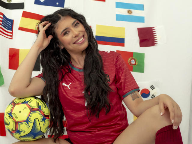 MiluSky with long black hair, smiling, wearing a red soccer shirt, sitting with a yellow and blue soccer ball, red socks, and flags behind her. Her tits are small.