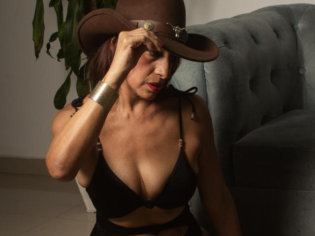 RebecaMonnique met een bruine hoed, een zwarte bh die haar borsten laat zien, een gouden armband, rode lipstick, die haar hoed aanraakt, zittend op een grijze bank, een plant op de achtergrond, rode lippen, sexy, volwassen..