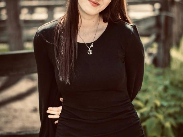 RuthaApe with long brown hair, wearing a black long-sleeve top, silver pentagram necklace, and hands on her hip, showing her small tits and subtle smile.