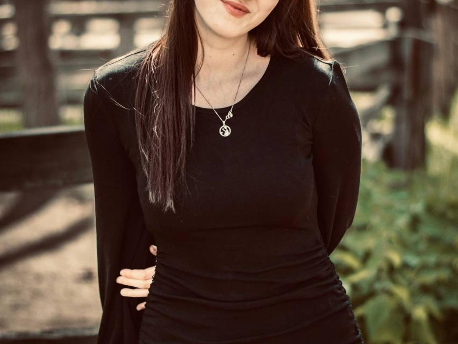 RuthaApe with long brown hair, wearing a black long-sleeve top, silver pentagram necklace, and hands on her hip, showing her small tits and subtle smile.