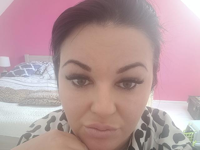 LiaLust has dark eyebrows, long eyelashes, and pouty lips, with black hair, wearing a patterned shirt, looking directly at the camera. Her hands rest on her chin.
