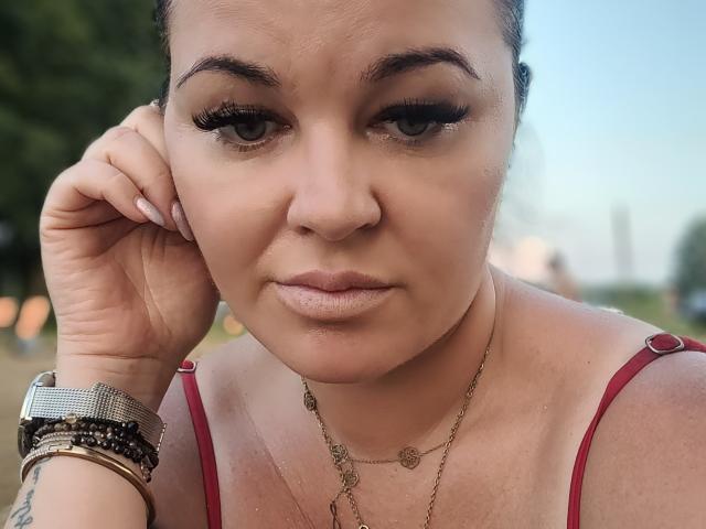 LiaLust heeft een serieus gezicht met lange wimpers, bruine ogen en een lichte huid. Ze draagt een rode top met bandjes, een gouden ketting en meerdere armbanden..