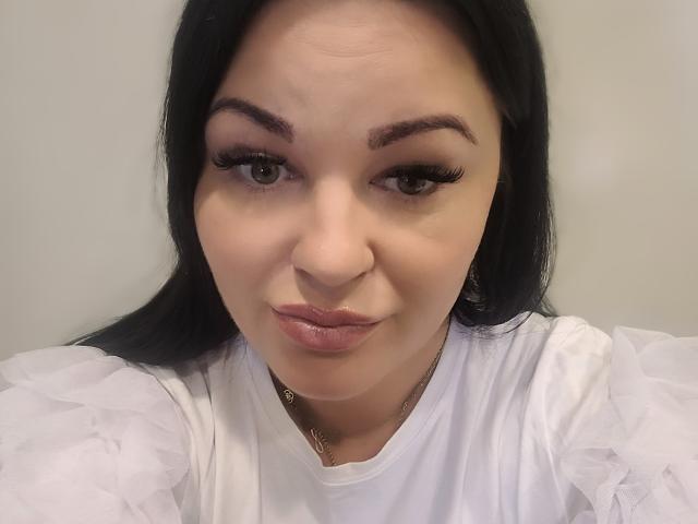 LiaLust, com cabelo preto, olhos castanhos e longos cílios, vestindo uma blusa branca com mangas bufantes, tem lábios rosados e um leve sorriso, olhando para a câmera..