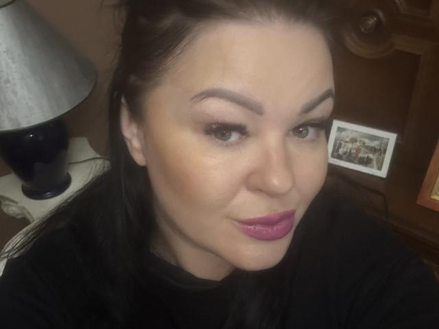 LiaLust has pale skin, dark brown hair, and pink lipstick. She's looking at the camera with a slight smile, wearing a black shirt. Her eyebrows are thick and arched.