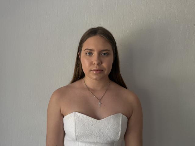 Margrett Munley, mit glattem, braunem Haar, trägt ein weißes, trägerloses Kleid, hat kleine Brüste, eine Kreuzkette und einen neutralen Gesichtsausdruck und steht vor einer schlichten Wand..