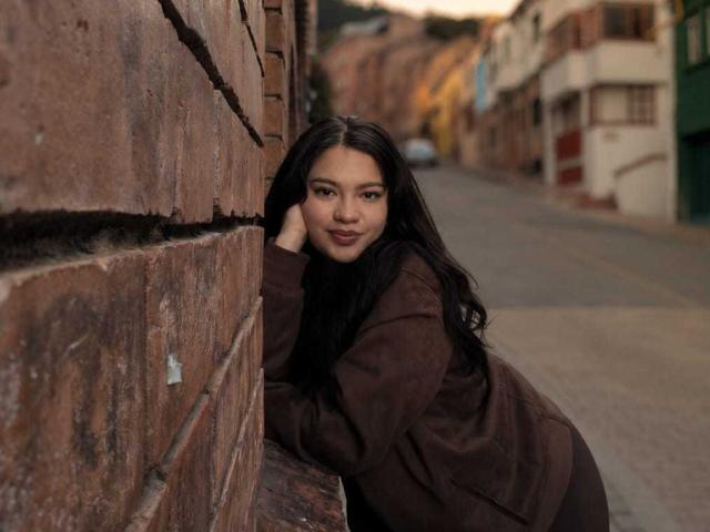 Elekktra with long black hair, brown jacket, leaning against a brick wall, smiling softly, her head resting on her hand, ass slightly raised, street in the background.