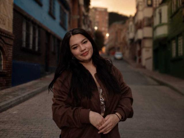 Elekktra with long black hair, brown jacket, and subtle smile, standing on a street, hands crossed, evening light, soft glow, slight red lipstick, calm and confident expression.
