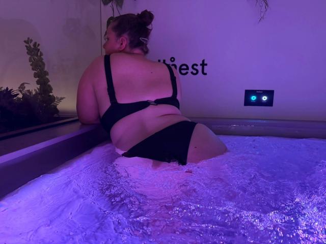 Sweety98, uma mulher sensual, plus size com uma bunda grande, vestindo um biquíni preto, inclinada em uma banheira de hidromassagem com luzes roxas, com água espirrando ao redor de sua bunda. Suas costas estão voltadas para a câmera..
