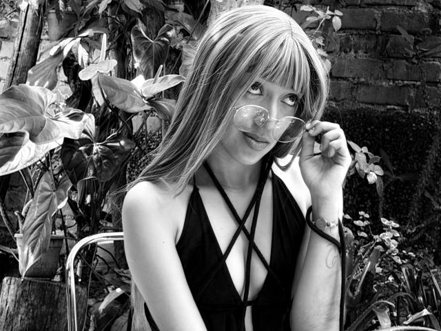 MichaelaMistress with long blonde hair, wearing a black strappy top, holding her round sunglasses, showing off her small tits, standing in front of leafy plants, in black and white.