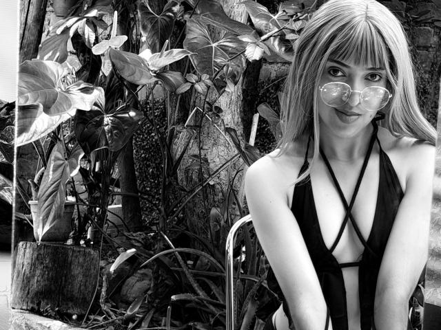 Black-and-white photo of MichaelaMistress with long blonde hair, wearing a strappy black bikini, and round sunglasses, leaning forward with a cute smile, showing her small tits.