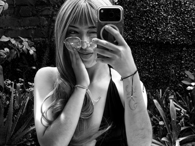Selfie in bianco e nero di MichaelaMistress con lunghi capelli biondi ondulati, che indossa occhiali, sorridente, con una mano sulla guancia, che tiene il telefono, con un tatuaggio visibile sul polso destro, che indossa un top a fascia..