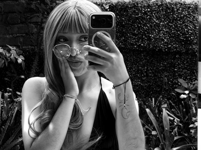 Schwarz-weißes Selfie von MichaelaMistress mit langen blonden Haaren, die ein dunkles Halstuch-Top trägt, ein Telefon hält, eine Brille auf der Nase trägt, die Zunge herausstreckt und ein Tattoo am rechten Arm hat..