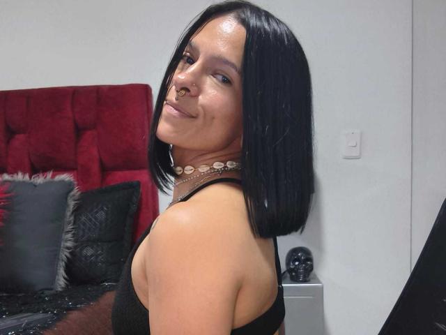 AmelieBlake with shiny black bob, smiling, wearing a black tank top, necklace, and nose ring, standing in a bedroom with a red tufted headboard and black pillows.